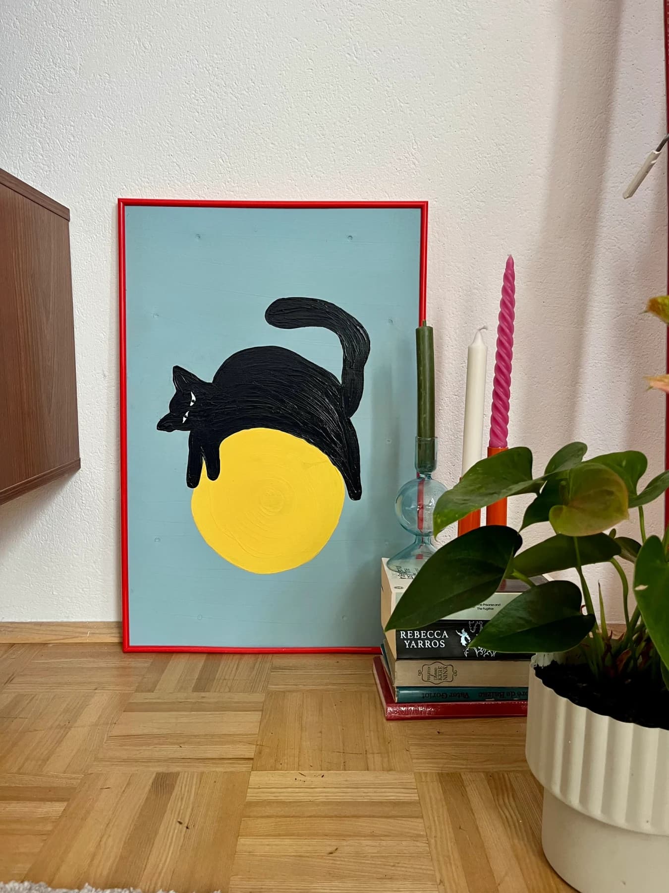 Gemälde „Muffy“ von Kristina Debertin im roten Rahmen, an eine Wand gelehnt: eine schwarze Katze liegt über einem gelben Kreis auf hellblauem Grund; daneben Kerzen, ein Stapel Bücher und eine Pflanze