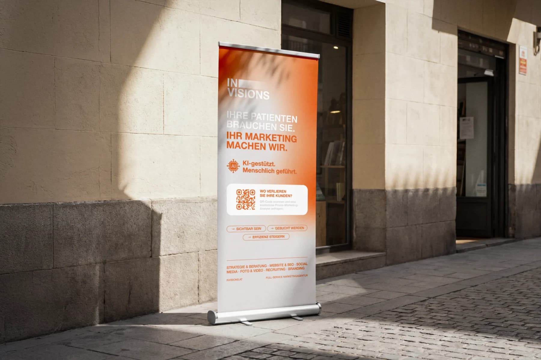 Roll-Up-Banner für die Marketingagentur Invisions, Mockup vor einer Hauswand in der Sonne: Verlauf von Orange zu Weiß, die Zeilen „Ihre Patienten brauchen Sie. Ihr Marketing machen wir.“, ein QR-Code und die Leistungen der Agentur